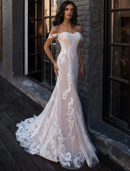 Bride in an elegant white lace mermaid wedding dress with off-shoulder cap sleeves and a court train, posing outdoors.